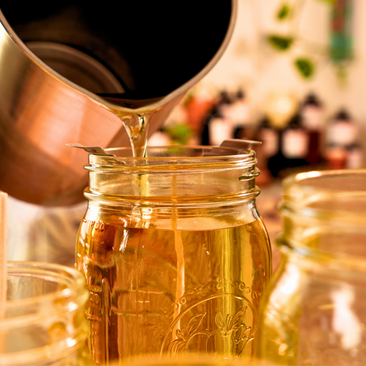 Pouring melted wax from a metal container into a glass jar with a blurred background of bottles.