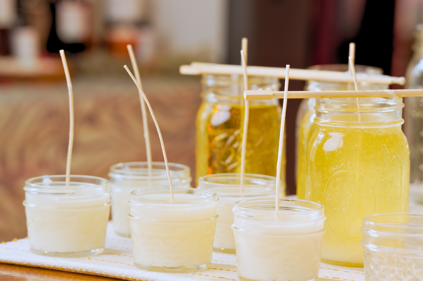 Small candles with tall wicks and jars of yellow liquid wax on a wooden surface.
