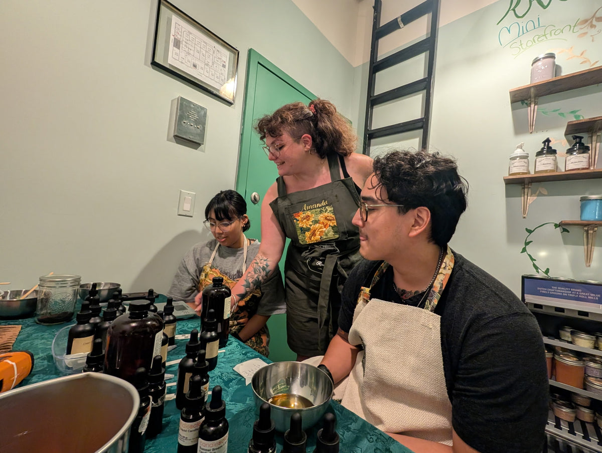 Three people wearing aprons gather around a table filled with small cosmetic or fragrance bottles in a workshop or studio setting. The room has light green walls, shelves with jars, and a green door in the background.